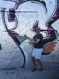 A man in a casual outfit, including a dark t-shirt, light checkered shorts, and sandals, is posing enthusiastically in front of a large piece of graffiti. The artwork is vibrant with silver, black, purple, orange, and teal colors forming abstract shapes on a wall.