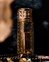 A decorative perfume bottle with intricate gold and brown patterns, placed against a dark background. Surrounding the bottle are beads and smoke, adding a luxurious and mysterious atmosphere.