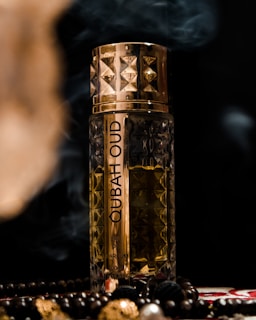 A decorative perfume bottle with intricate gold and brown patterns, placed against a dark background. Surrounding the bottle are beads and smoke, adding a luxurious and mysterious atmosphere.