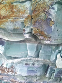Layers of sedimentary rock are visible, with various shades of green and some rusty brown coloration. The surface of the rocks is uneven and cracked, with distinct layers that suggest geological formations.