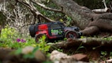 A toy off-road vehicle with vibrant red and black colors is positioned on a natural terrain with green foliage and a large twisted tree in the background. The ground is covered with grass and small purple flowers.