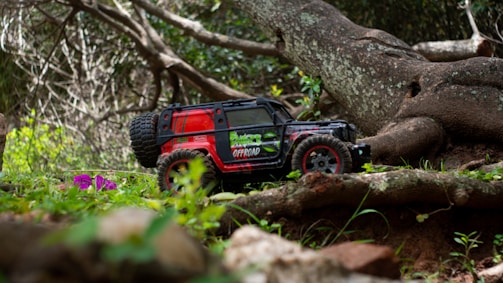A toy off-road vehicle with vibrant red and black colors is positioned on a natural terrain with green foliage and a large twisted tree in the background. The ground is covered with grass and small purple flowers.