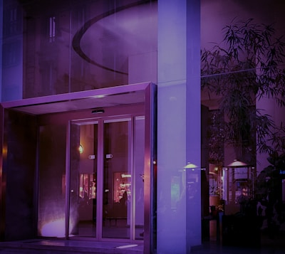 A modern building entrance featuring glass doors and illuminated interior lighting. The reflections on the glass surfaces create a slightly abstract appearance, with visible plants and interior decor elements. The color palette is dominated by purple and blue hues, casting a serene and calm ambiance.