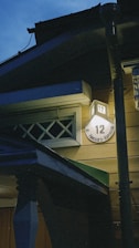 A nighttime view of a building corner illuminated by a light. A house number '12' is clearly visible on a round sign attached to the wall, with some text beneath it. The architecture features wooden siding and a small overhang or awning. The sky is a deep blue, indicating evening or early dawn.