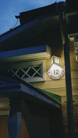 A nighttime view of a building corner illuminated by a light. A house number '12' is clearly visible on a round sign attached to the wall, with some text beneath it. The architecture features wooden siding and a small overhang or awning. The sky is a deep blue, indicating evening or early dawn.