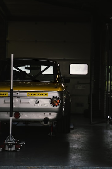 A vintage BMW 2002 car with a yellow and white color scheme is parked inside a dimly lit garage. The car is partially elevated using a jack stand, and a Dunlop logo is prominently displayed on its rear.