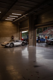 A dimly lit garage space with a sleek, vintage silver sports car parked near the entrance. The garage has concrete walls and a smooth, reflective floor. Several people are standing outside, near other cars, under an overcast sky.