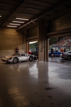 A dimly lit garage space with a sleek, vintage silver sports car parked near the entrance. The garage has concrete walls and a smooth, reflective floor. Several people are standing outside, near other cars, under an overcast sky.