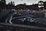 A car race track surrounded by spectators, multiple cars are seen performing drifts on the asphalt. Several people in high-visibility vests are positioned around the area, possibly as event staff. Various banners and advertisements are displayed along the track, with forested hills in the background.