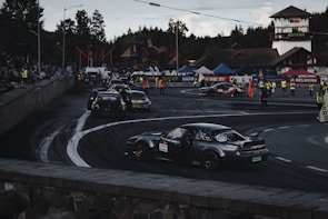 A car race track surrounded by spectators, multiple cars are seen performing drifts on the asphalt. Several people in high-visibility vests are positioned around the area, possibly as event staff. Various banners and advertisements are displayed along the track, with forested hills in the background.