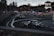 A car race track surrounded by spectators, multiple cars are seen performing drifts on the asphalt. Several people in high-visibility vests are positioned around the area, possibly as event staff. Various banners and advertisements are displayed along the track, with forested hills in the background.