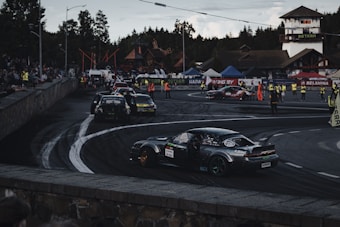 A car race track surrounded by spectators, multiple cars are seen performing drifts on the asphalt. Several people in high-visibility vests are positioned around the area, possibly as event staff. Various banners and advertisements are displayed along the track, with forested hills in the background.