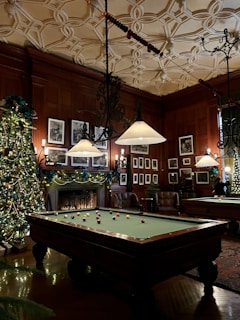 A luxurious, warmly lit room featuring an ornately decorated ceiling and wood-paneled walls adorned with framed photographs. A billiards table takes center stage, surrounded by holiday decorations including a large, ornament-laden Christmas tree. Comfortable leather armchairs are placed near a fireplace, contributing to the cozy atmosphere.
