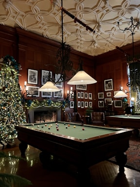 A luxurious, warmly lit room featuring an ornately decorated ceiling and wood-paneled walls adorned with framed photographs. A billiards table takes center stage, surrounded by holiday decorations including a large, ornament-laden Christmas tree. Comfortable leather armchairs are placed near a fireplace, contributing to the cozy atmosphere.