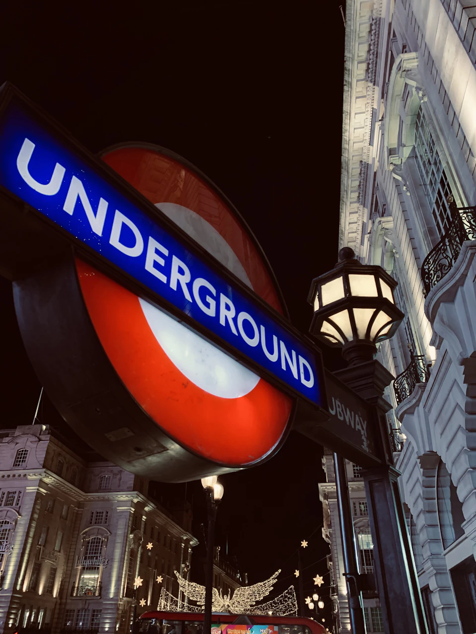 The image shows a street scene at night, with a prominent Underground sign in the foreground. The sign is a large circular structure painted in red and white colors, featuring an illuminated white "Underground" logo on top. Below the logo, there's a smaller red circle with a white border that reads "Underground". The sign is situated next to a street lamp, which casts a warm glow onto the scene. In the background, there are buildings lining the street, and a few cars can be seen driving by.