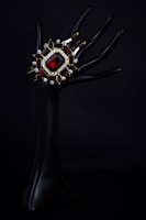 An elegant dark mannequin hand holds a luxurious bracelet adorned with a large red gemstone, surrounded by smaller gemstones and intricate gold settings. The background is pitch black, highlighting the jewelry's vivid colors and intricate design.