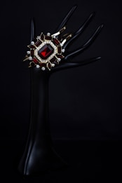 An elegant dark mannequin hand holds a luxurious bracelet adorned with a large red gemstone, surrounded by smaller gemstones and intricate gold settings. The background is pitch black, highlighting the jewelry's vivid colors and intricate design.