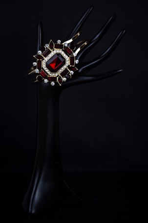 An elegant dark mannequin hand holds a luxurious bracelet adorned with a large red gemstone, surrounded by smaller gemstones and intricate gold settings. The background is pitch black, highlighting the jewelry's vivid colors and intricate design.