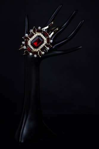An elegant dark mannequin hand holds a luxurious bracelet adorned with a large red gemstone, surrounded by smaller gemstones and intricate gold settings. The background is pitch black, highlighting the jewelry's vivid colors and intricate design.