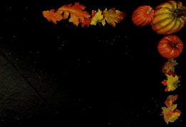 A dark background adorned with an arrangement of orange pumpkins and autumn leaves. The leaves, in shades of orange, yellow, and red, are positioned in the top left and right corners, while the pumpkins are clustered at the top right.