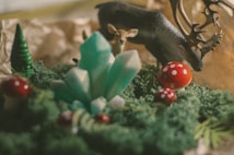 A whimsical scene featuring a miniature forest setting with vibrant green moss and red mushrooms with white spots. In the foreground, there are several turquoise crystals emerging from the moss. A small deer figurine and a stag with antlers are positioned in the background, creating an enchanting woodland atmosphere.