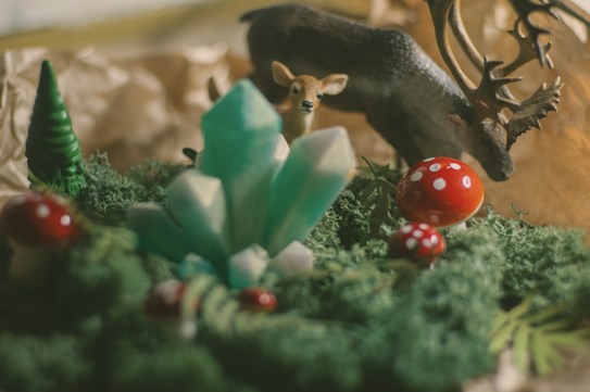 A whimsical scene featuring a miniature forest setting with vibrant green moss and red mushrooms with white spots. In the foreground, there are several turquoise crystals emerging from the moss. A small deer figurine and a stag with antlers are positioned in the background, creating an enchanting woodland atmosphere.