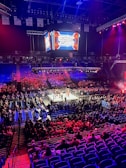 An indoor arena is filled with people watching a wrestling match. The ring is centrally located, and surrounded by audience members sitting in tiered seating. The arena is illuminated with red and blue lights, and a large overhead screen displays 'Sunday Stunner.' Spectators are engrossed in the match taking place in the well-lit ring.