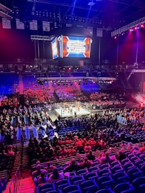 An indoor arena is filled with people watching a wrestling match. The ring is centrally located, and surrounded by audience members sitting in tiered seating. The arena is illuminated with red and blue lights, and a large overhead screen displays 'Sunday Stunner.' Spectators are engrossed in the match taking place in the well-lit ring.