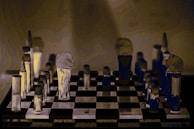 A stylized image of a chessboard with vertically elongated, abstract chess pieces. The pieces have an artistic, distorted appearance with a mix of dark and light tones that create a surreal atmosphere. The background is muted, focusing attention on the chessboard and its unique pieces.