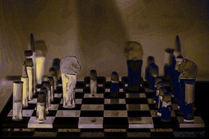 A stylized image of a chessboard with vertically elongated, abstract chess pieces. The pieces have an artistic, distorted appearance with a mix of dark and light tones that create a surreal atmosphere. The background is muted, focusing attention on the chessboard and its unique pieces.