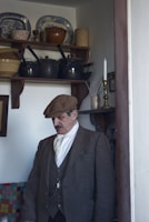 A man wearing a vintage-style suit and a flat cap stands in a room with antique kitchen items. Shelves behind him hold various pottery and metal pots. A single lit candle is on a nearby shelf, adding a historical ambiance.
