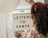 A child with curly hair tied with a red velvet bow is holding a white balloon and placing a letter into a mailbox labeled 'Letters to Santa.' The child is wearing a colorful floral-patterned dress.