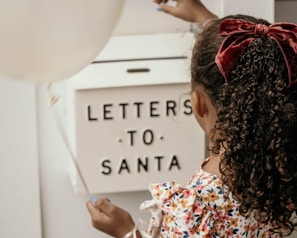 A child with curly hair tied with a red velvet bow is holding a white balloon and placing a letter into a mailbox labeled 'Letters to Santa.' The child is wearing a colorful floral-patterned dress.