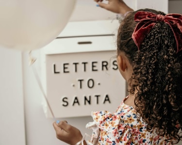 A child with curly hair tied with a red velvet bow is holding a white balloon and placing a letter into a mailbox labeled 'Letters to Santa.' The child is wearing a colorful floral-patterned dress.