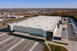 Aerial view of a large warehouse facility with a white, flat roof surrounded by extensive parking areas. The structure features multiple loading docks along one side. It is situated near a commercial area and farmland, with a clear sky visible above.