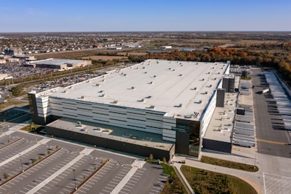 Aerial view of a large warehouse facility with a white, flat roof surrounded by extensive parking areas. The structure features multiple loading docks along one side. It is situated near a commercial area and farmland, with a clear sky visible above.