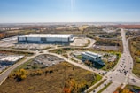 Aerial view of a large industrial complex with a warehouse or building surrounded by a parking lot. The area is connected by several roads, including a major highway, with vehicles visible on them. The landscape features some greenery with trees showing autumn colors. Residential and commercial buildings are seen in the background, with vast open fields extending into the horizon.