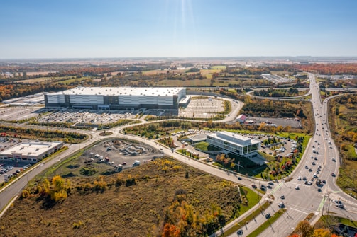 Aerial view of a large industrial complex with a warehouse or building surrounded by a parking lot. The area is connected by several roads, including a major highway, with vehicles visible on them. The landscape features some greenery with trees showing autumn colors. Residential and commercial buildings are seen in the background, with vast open fields extending into the horizon.