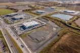 Aerial view of a commercial area surrounded by open fields and a large pond. Several modern buildings with parking lots are visible, along with a network of roads and cars. The area is bordered by trees and farmland, showcasing a blend of urban and rural landscapes.