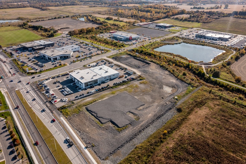 Aerial view of a commercial area surrounded by open fields and a large pond. Several modern buildings with parking lots are visible, along with a network of roads and cars. The area is bordered by trees and farmland, showcasing a blend of urban and rural landscapes.
