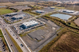 Aerial view of a commercial area surrounded by open fields and a large pond. Several modern buildings with parking lots are visible, along with a network of roads and cars. The area is bordered by trees and farmland, showcasing a blend of urban and rural landscapes.