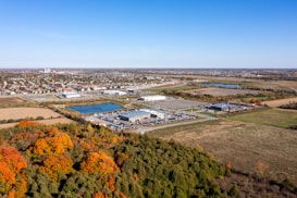 Aerial view of a suburban area featuring a mix of industrial buildings, parking lots, and residential neighborhoods in the distance. Autumn foliage with vibrant orange and green tones is visible in the foreground alongside a couple of ponds.