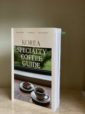 A book titled 'Korea Specialty Coffee Guide' is standing upright on a marble surface. The cover features an image of a cup of coffee and a piece of dessert on a wooden table set against a window with greenery outside.