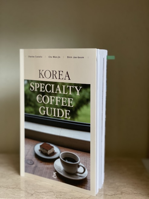 A book titled 'Korea Specialty Coffee Guide' is standing upright on a marble surface. The cover features an image of a cup of coffee and a piece of dessert on a wooden table set against a window with greenery outside.