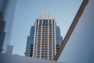 A high-rise building with a modern architectural design featuring a symmetrical pattern of balconies and blue reflective glass windows. The top of the building has a distinctive pointed structure and it is framed by geometric angles of nearby structures.