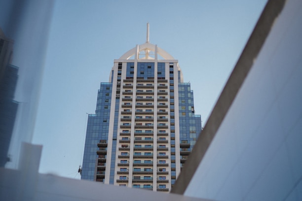 A high-rise building with a modern architectural design featuring a symmetrical pattern of balconies and blue reflective glass windows. The top of the building has a distinctive pointed structure and it is framed by geometric angles of nearby structures.