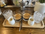 A wooden tray holds two glasses of espresso, two small white pitchers, and two glasses filled with ice cubes. The setup is arranged neatly on a wooden table, creating a rustic and cozy atmosphere. In the background, part of a moka pot and a potted plant are visible, suggesting a coffee setting.