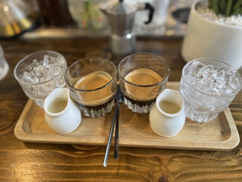 A wooden tray holds two glasses of espresso, two small white pitchers, and two glasses filled with ice cubes. The setup is arranged neatly on a wooden table, creating a rustic and cozy atmosphere. In the background, part of a moka pot and a potted plant are visible, suggesting a coffee setting.