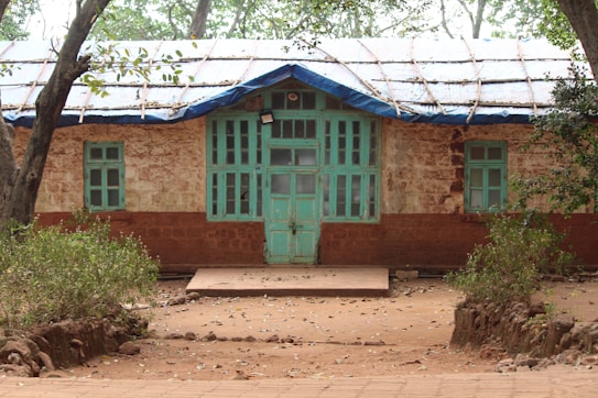 A rustic, single-story building with a thatched roof and wooden windows painted in turquoise. The structure is surrounded by trees, with patches of greenery on both sides of a dirt pathway leading to the entrance. The walls are a mix of exposed brick and plaster.