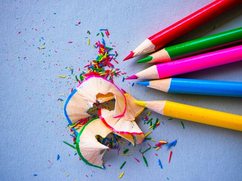 A collection of five vibrant colored pencils in shades of red, green, blue, pink, and yellow is arranged diagonally on a textured light gray background. In the foreground, pencil shavings and small specks of colorful pencil debris are scattered around, creating a playful and artistic scene.